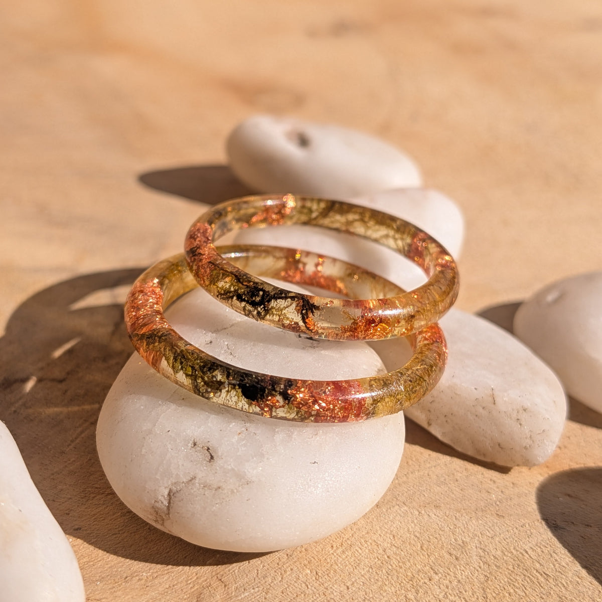 Two delicate resin rings with moss and copper flakes overlapping on a white pebble in sunlight, EL-Creations
