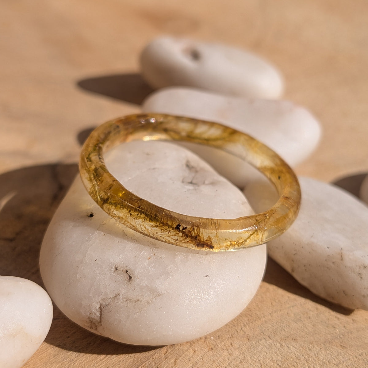 Slim woodland moss resin ring resting at an angle on white pebbles, showing botanical details inside the glossy band, EL-Creations