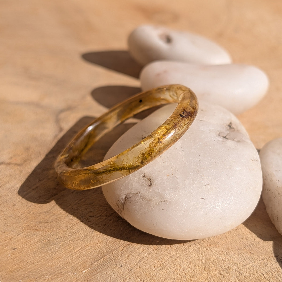 Side view of a slender moss resin ring leaning against a white pebble in sunlight, EL-Creations
