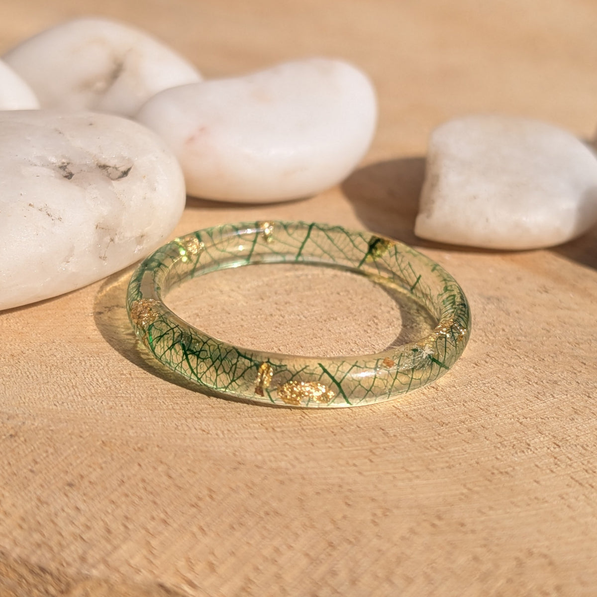 Thin resin ring with forest-green skeleton leaf veins and gold flakes lying flat on wood beside white pebbles, EL-Creations