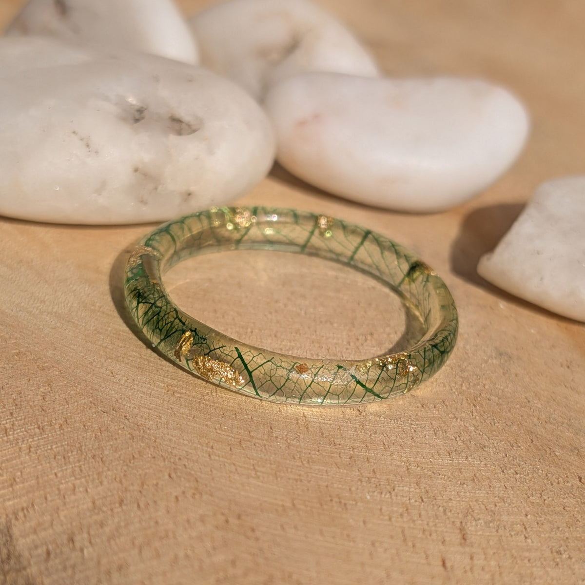 Angled view of a slim skeleton leaf resin ring with green veins and gold flakes lying on a wooden surface, EL-Creations