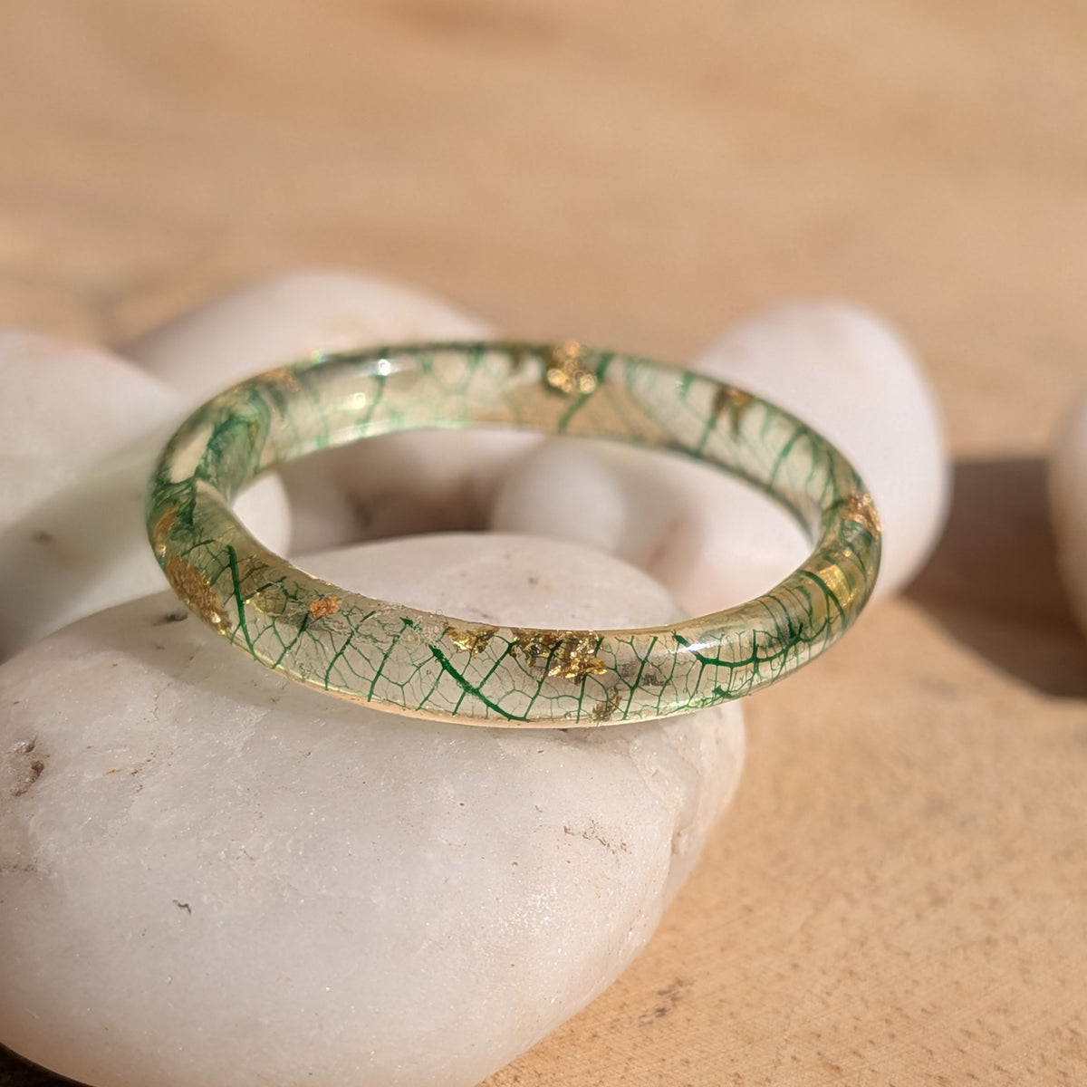 Close-up of a delicate resin ring with forest-green skeleton leaf veins and gold flakes displayed on white pebbles, EL-Creations