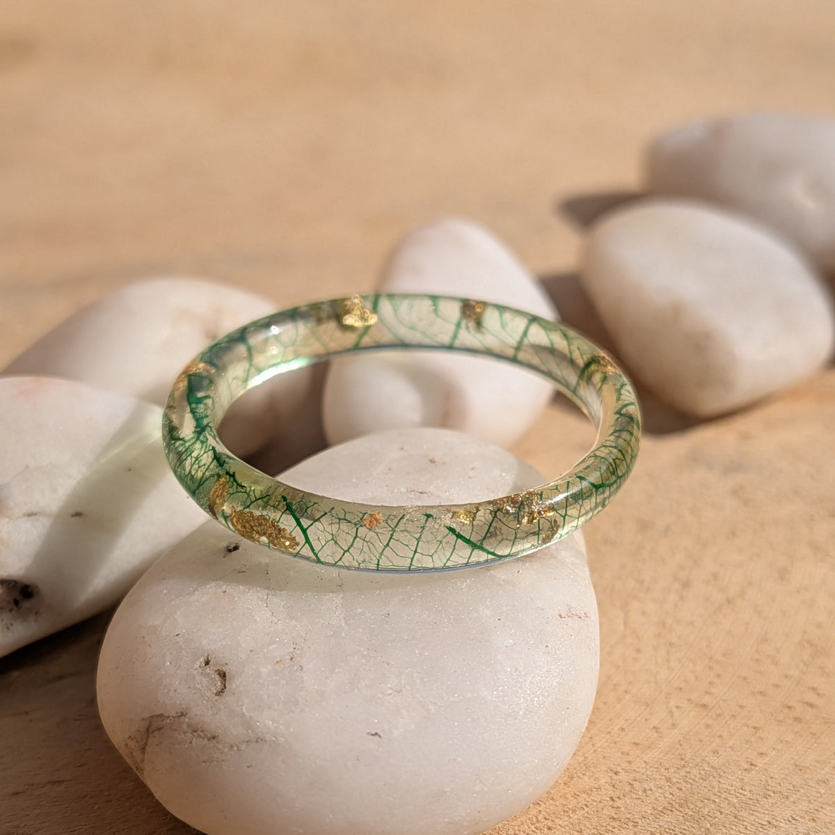 Close-up of a delicate resin ring with forest-green skeleton leaf veins and gold flakes displayed on white pebbles, EL-Creations
