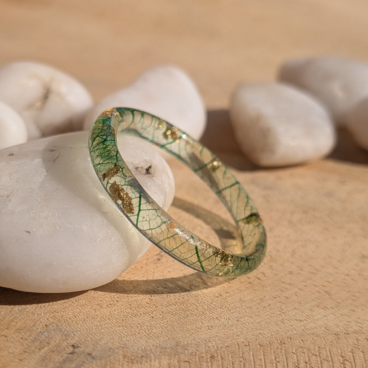 Forest-green skeleton leaf resin ring with gold flakes leaning upright against a white pebble in sunlight, EL-Creations