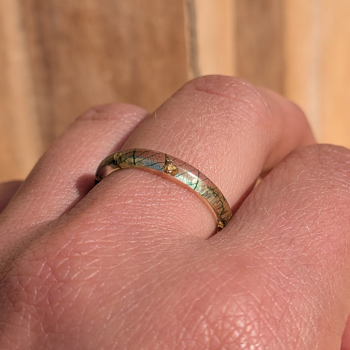Front close-up of a skeleton leaf resin ring worn on a finger, showing fine green veins and gold flakes beneath the glossy surface, EL-Creations