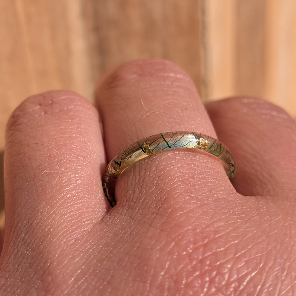 Front close-up of a skeleton leaf resin ring worn on a finger, showing fine green veins and gold flakes beneath the glossy surface, EL-Creations