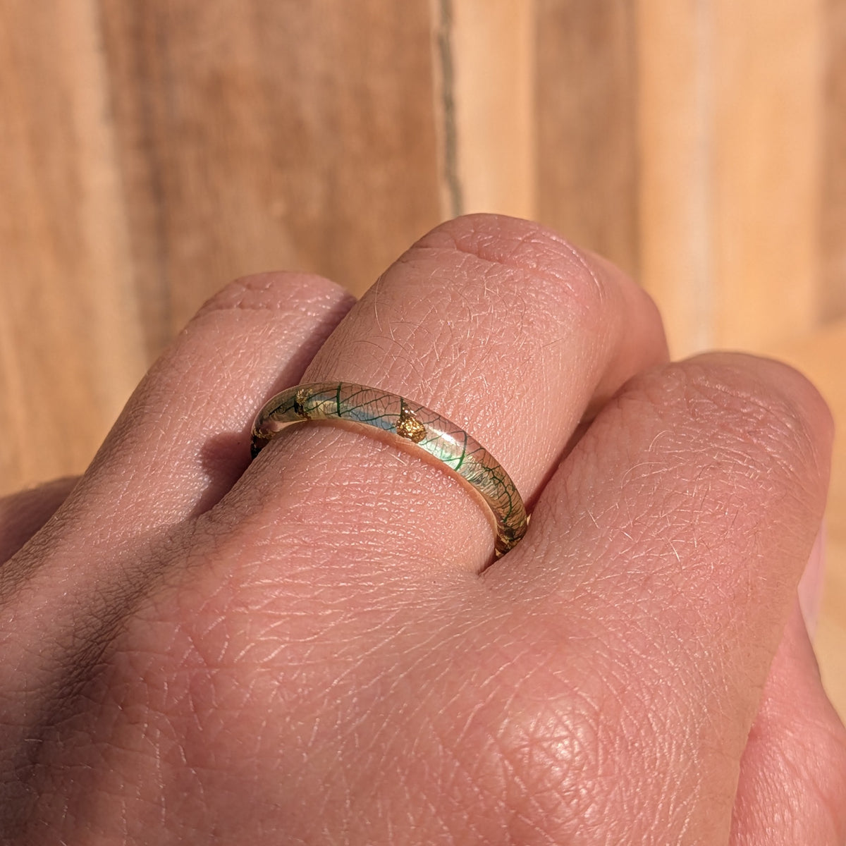 Slim botanical resin ring with forest-green leaf veins and gold flakes worn on a gently bent hand in sunlight, EL-Creations