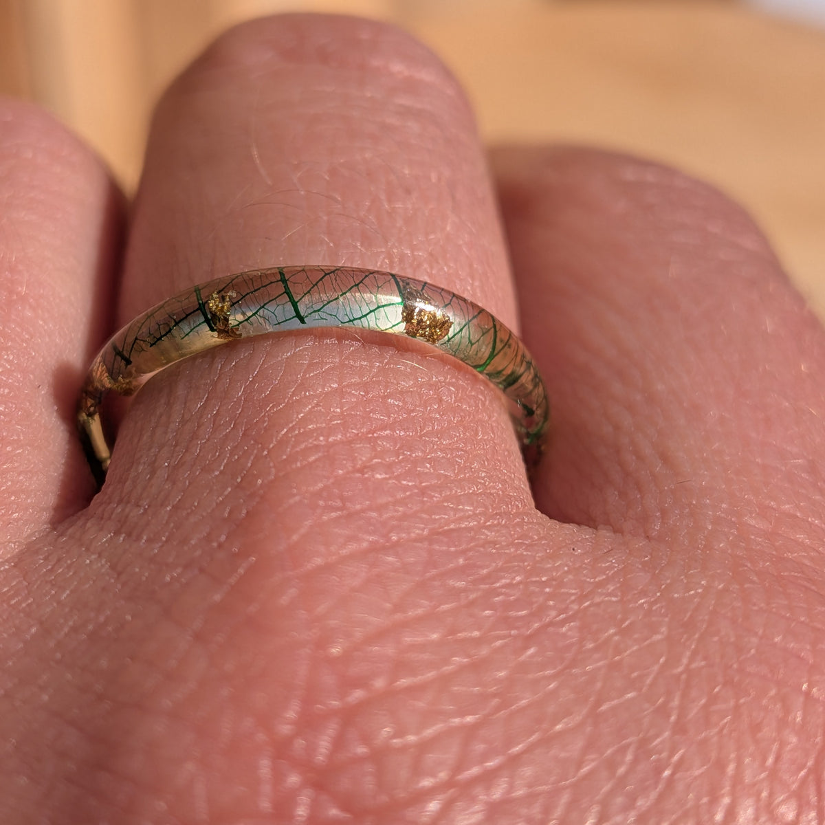 Macro close-up of forest-green skeleton leaf veins and gold flakes inside a thin resin ring worn on a finger, EL-Creations
