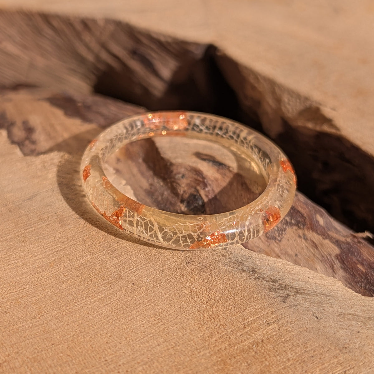 Angled view of a transparent resin ring with white leaf veins and copper flakes resting beside a hollow in the wood, EL-Creations