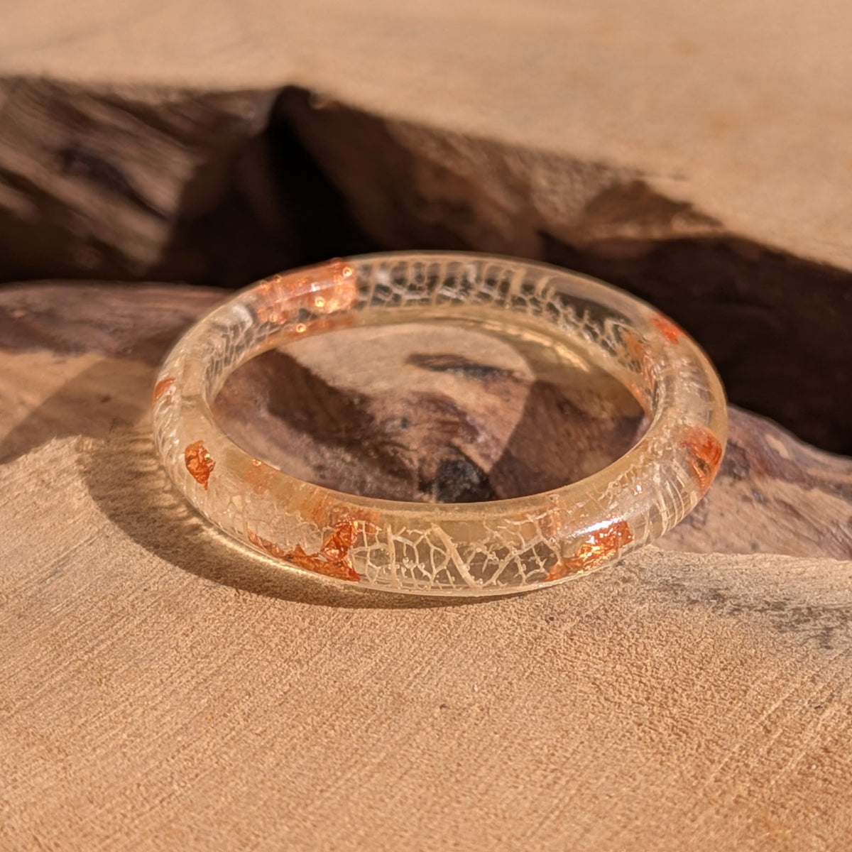 Thin resin ring with white skeleton leaf veins and copper flakes resting on textured wood beside a dark hollow, EL-Creations