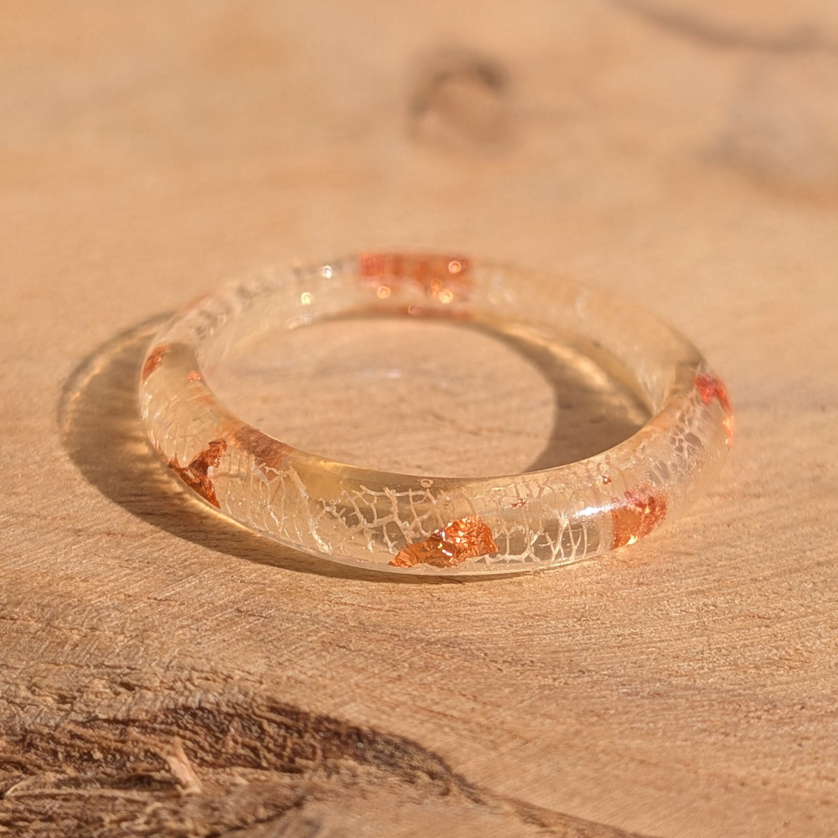 Close-up of a delicate white skeleton leaf resin ring with copper flakes lying flat on a sunlit wooden surface, EL-Creations