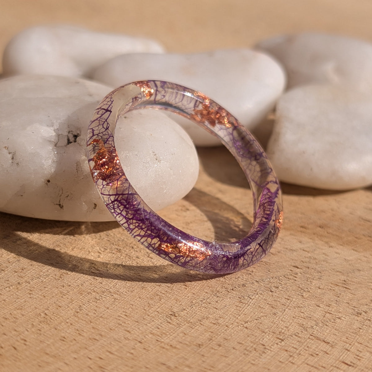 Purple skeleton leaf resin ring with copper flakes standing upright against a white pebble on a wooden surface, EL-Creations