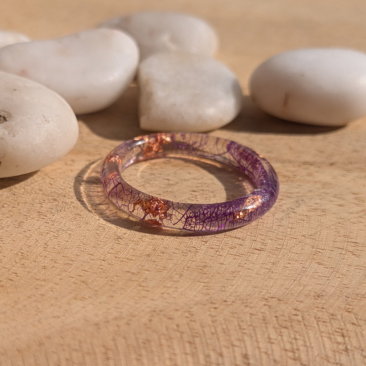 Thin transparent resin ring with purple leaf veins and copper flakes lying flat on wood beside white stones, EL-Creations