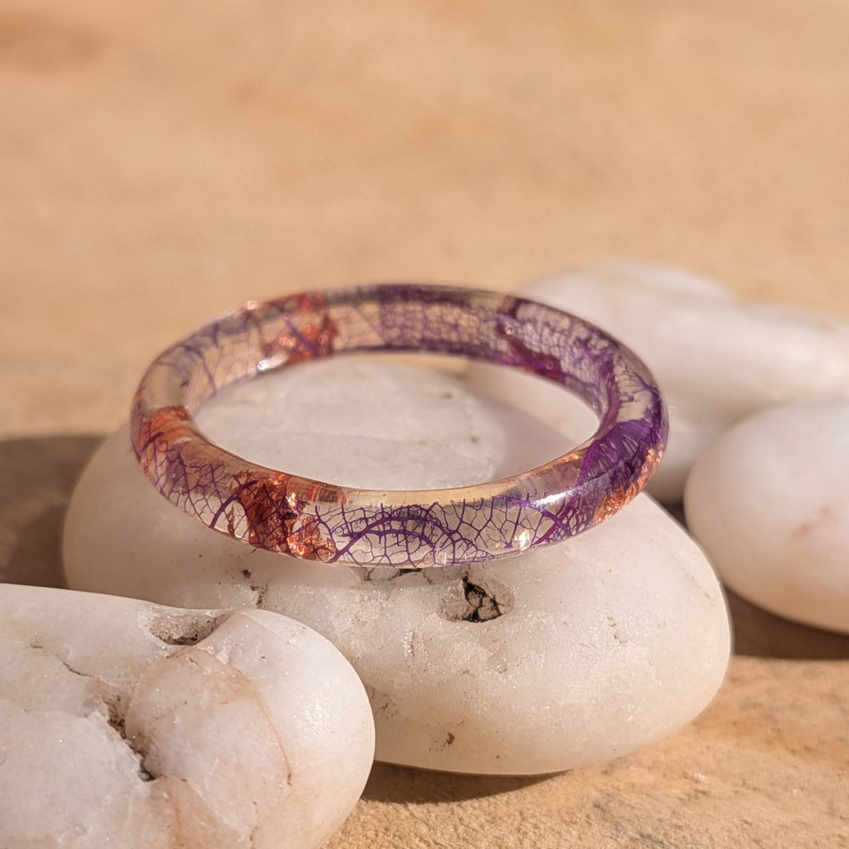 Delicate resin ring with purple skeleton leaf veins and copper flakes resting on white pebbles in sunlight, EL-Creations