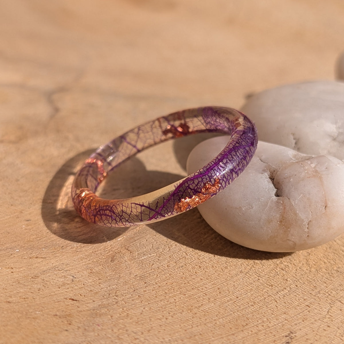 Angled side view of a slim purple skeleton leaf resin ring with copper flakes leaning against a white stone, EL-Creations