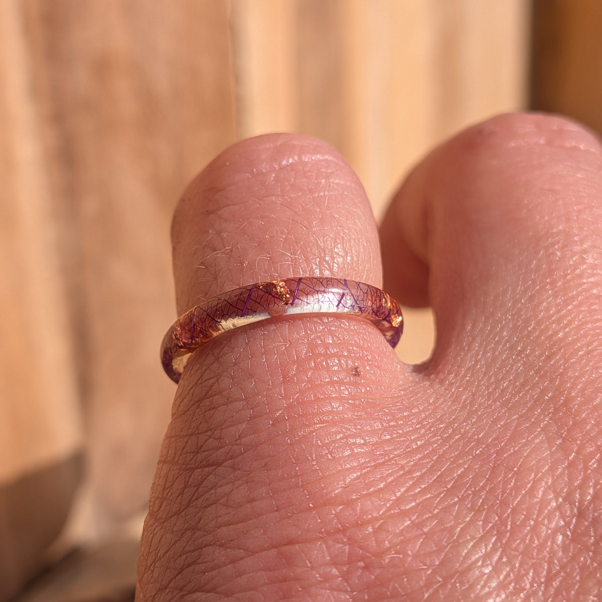 Front macro view of a thin resin ring worn on a finger, showing purple leaf veins and copper flakes beneath the glossy surface, EL-Creations