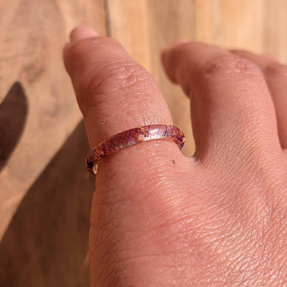 Thin botanical resin ring with purple leaf veins and copper flakes worn on a bent hand against a sunlit wooden background, EL-Creations