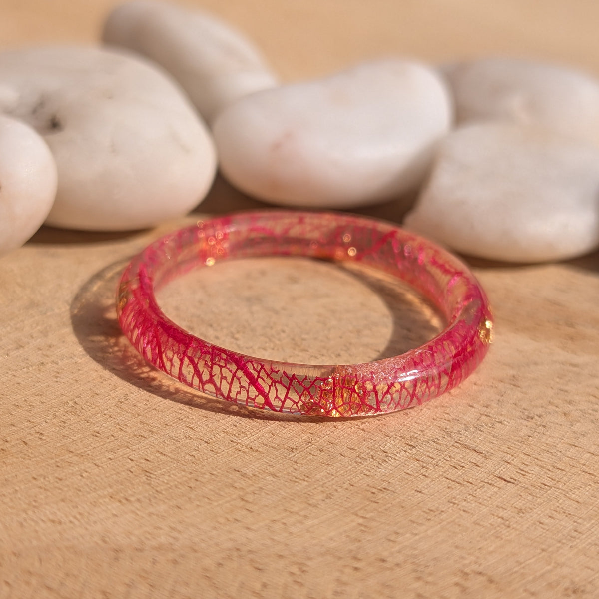 Thin transparent resin ring with red skeleton leaf veins and copper flakes lying flat on wood beside white pebbles, EL-Creations