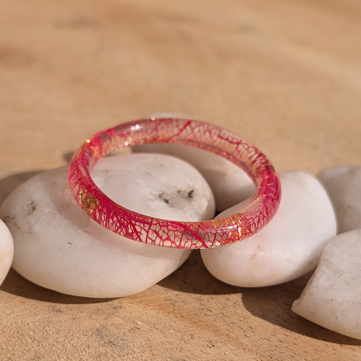Close-up of a thin botanical resin ring with red leaf veins and copper flakes displayed on white pebbles, EL-Creations