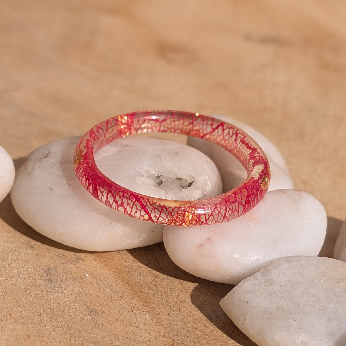 Red skeleton leaf resin ring with copper flakes resting across white stones in sunlight, showing its delicate rounded band, EL-Creations