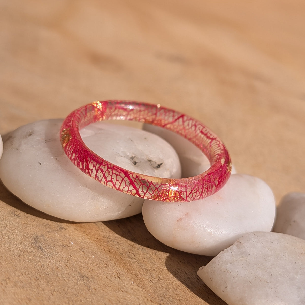 Delicate red skeleton leaf resin ring resting at an angle across white stones, with copper flakes visible inside the band, EL-Creations