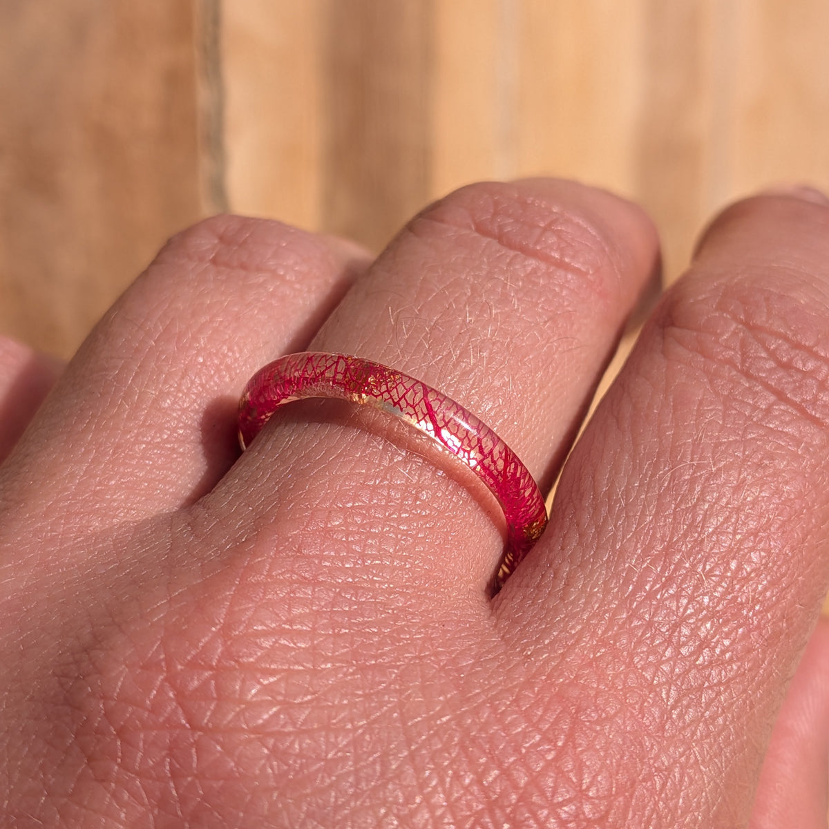 Angled close-up of a thin resin ring with red skeleton leaf veins and copper flakes worn on a finger in sunlight, EL-Creations
