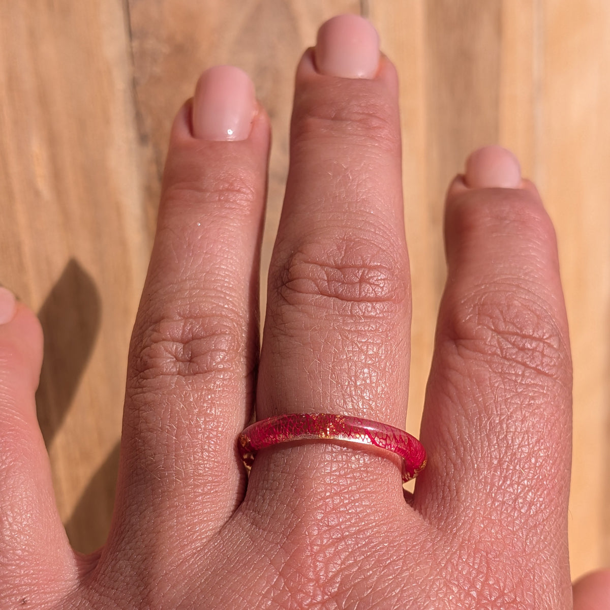 Thin botanical resin ring with red leaf veins and copper flakes worn on an outstretched hand against a wooden background, EL-Creations