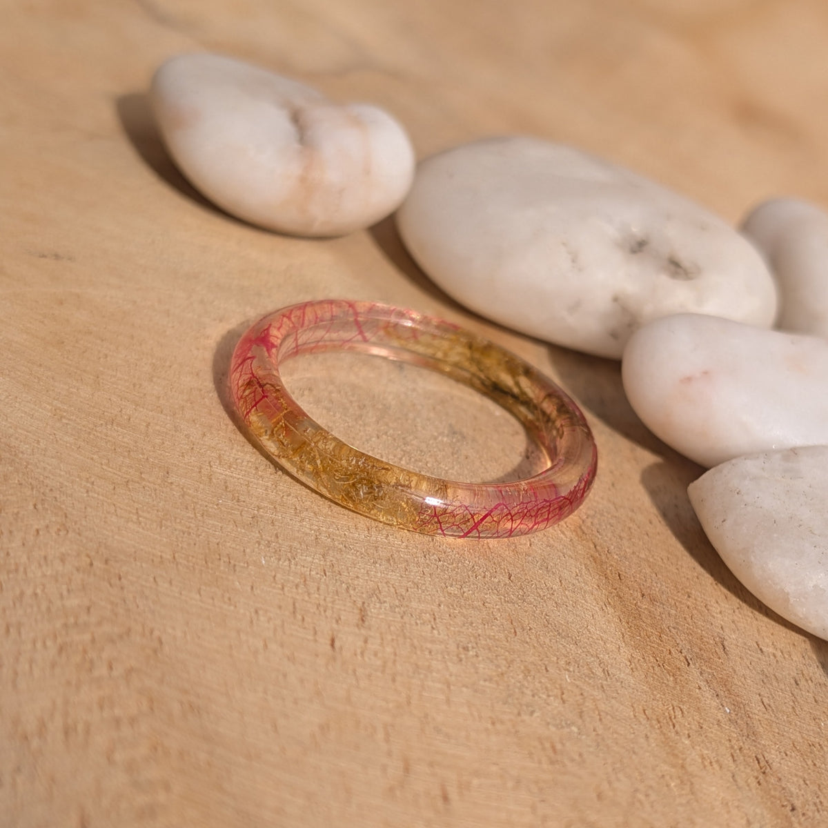 Angled view of a delicate resin ring with pink leaf veins and moss on a wooden surface beside white pebbles, EL-Creations
