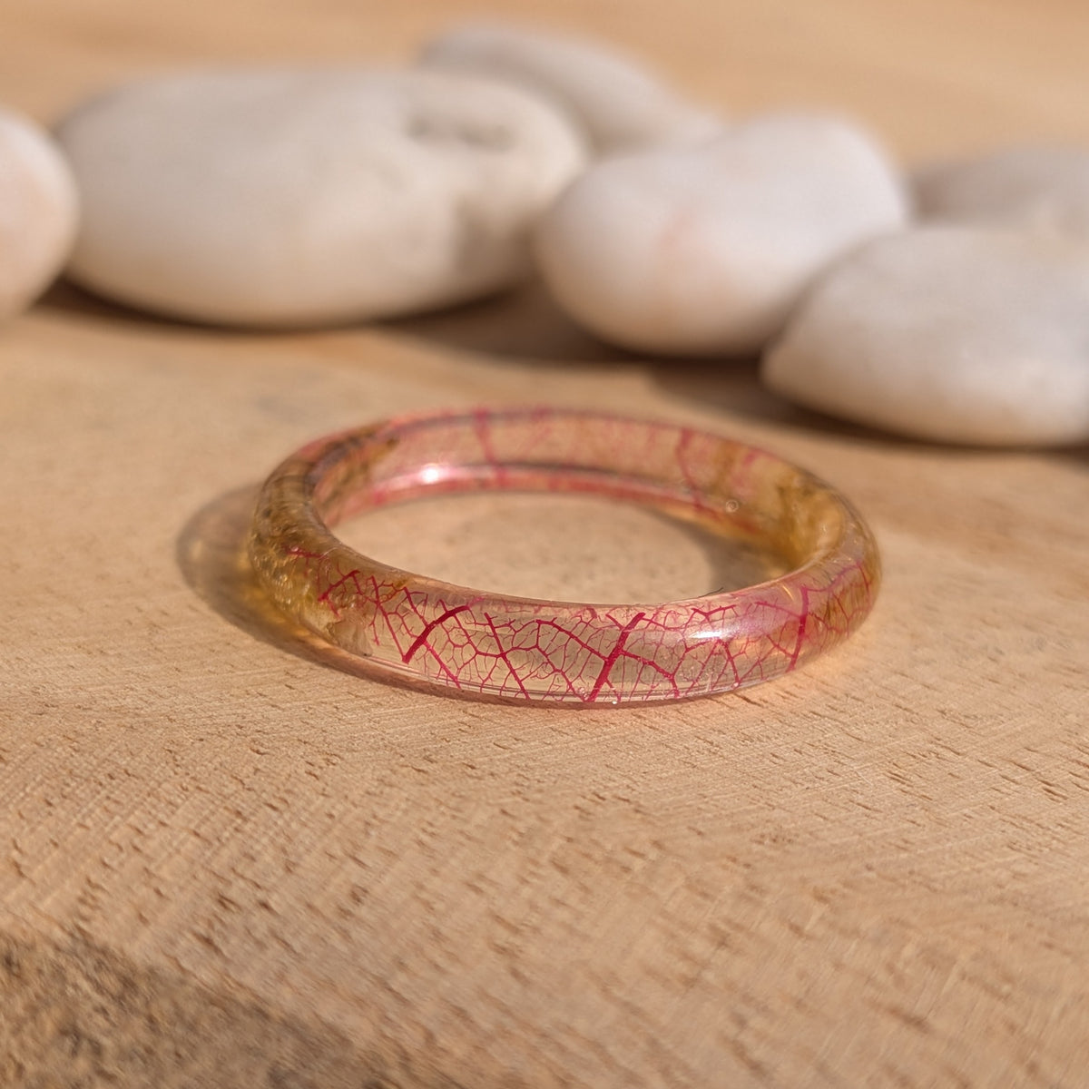 Close-up of pink skeleton leaf veins inside a thin glossy resin band lying on sunlit wood, EL-Creations
