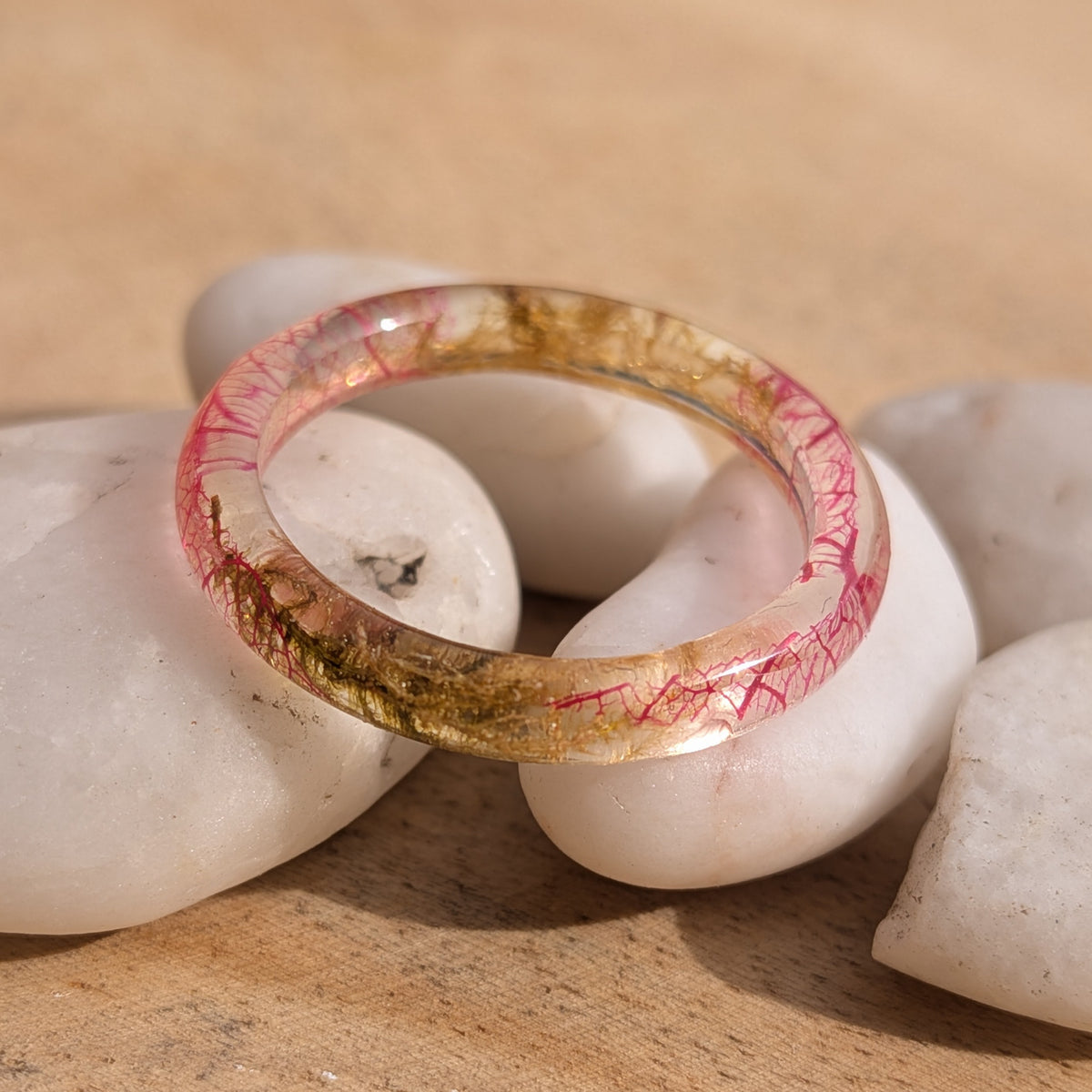 Thin transparent resin ring with pink skeleton leaf veins and moss resting across white pebbles in sunlight, EL-Creations
