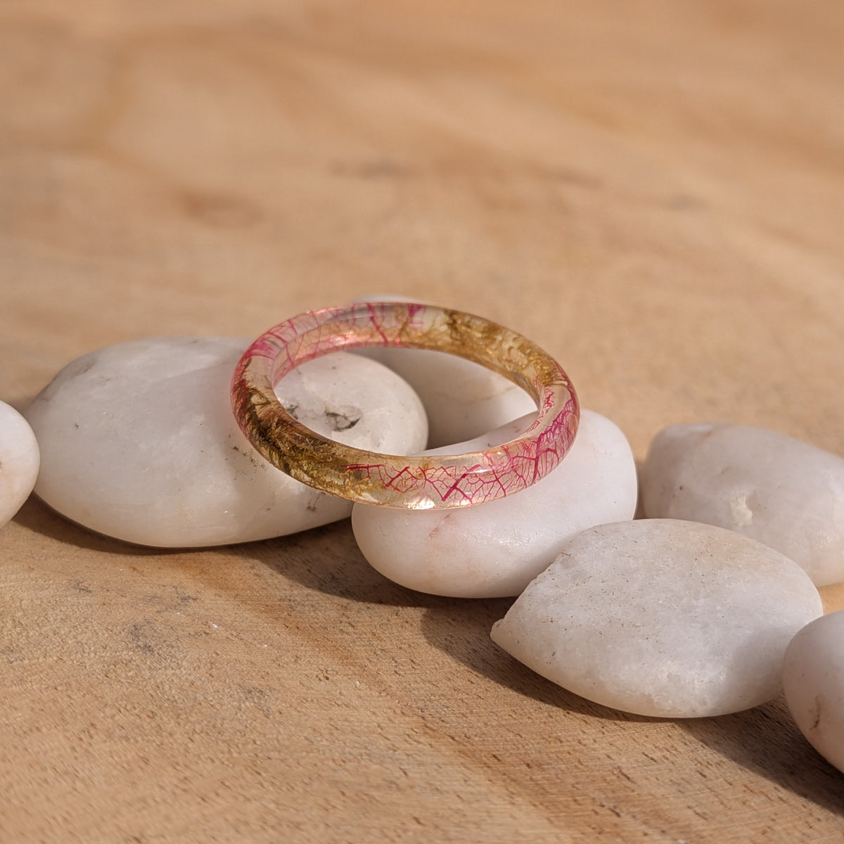 Thin pink leaf and moss resin ring displayed on white pebbles in a wider sunlit wooden setting, EL-Creations