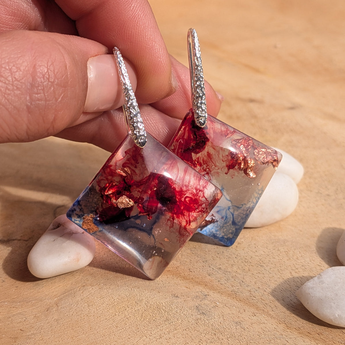 Handheld pair of crimson and blue resin earrings beside white stones, EL-Creations