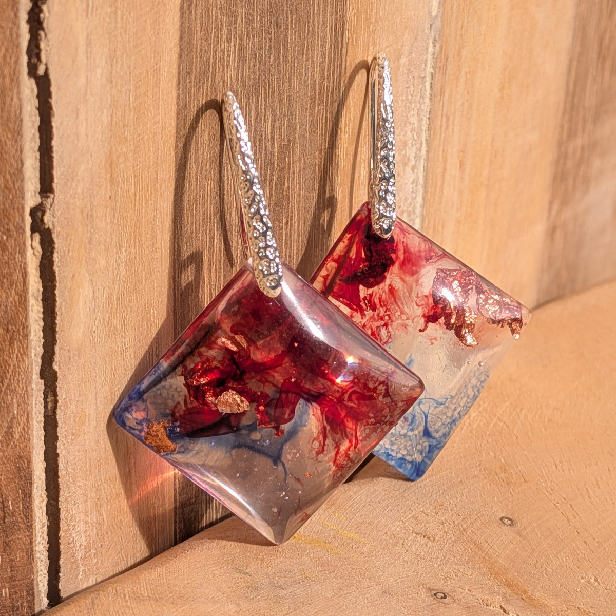 Square resin earrings with red and blue swirls hanging against wood, EL-Creations