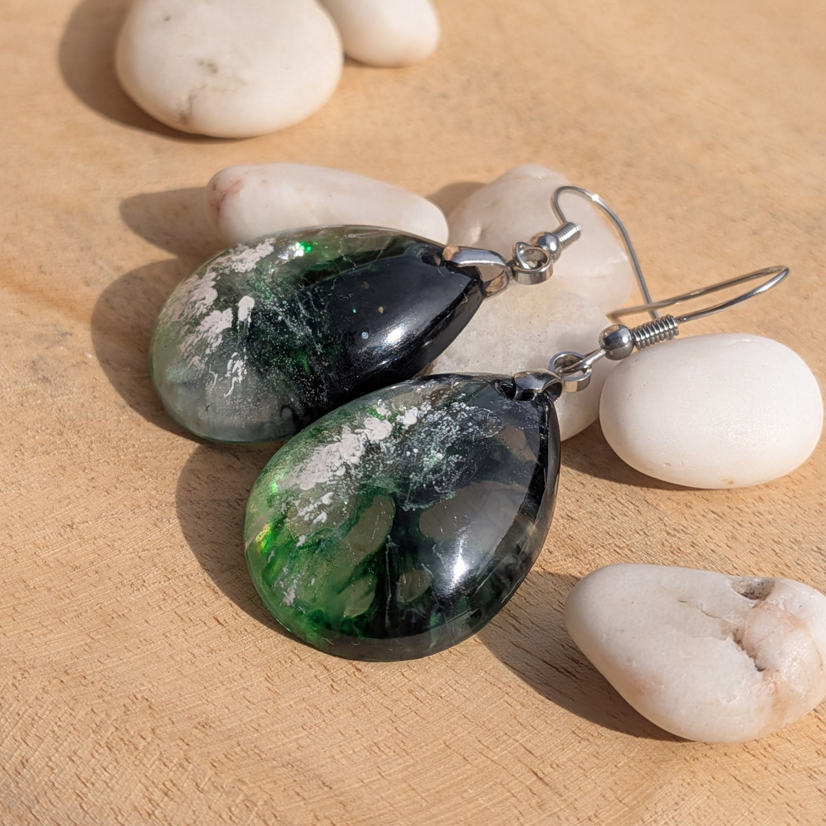 Green black resin earrings laid at an angle among white stones, EL-Creations