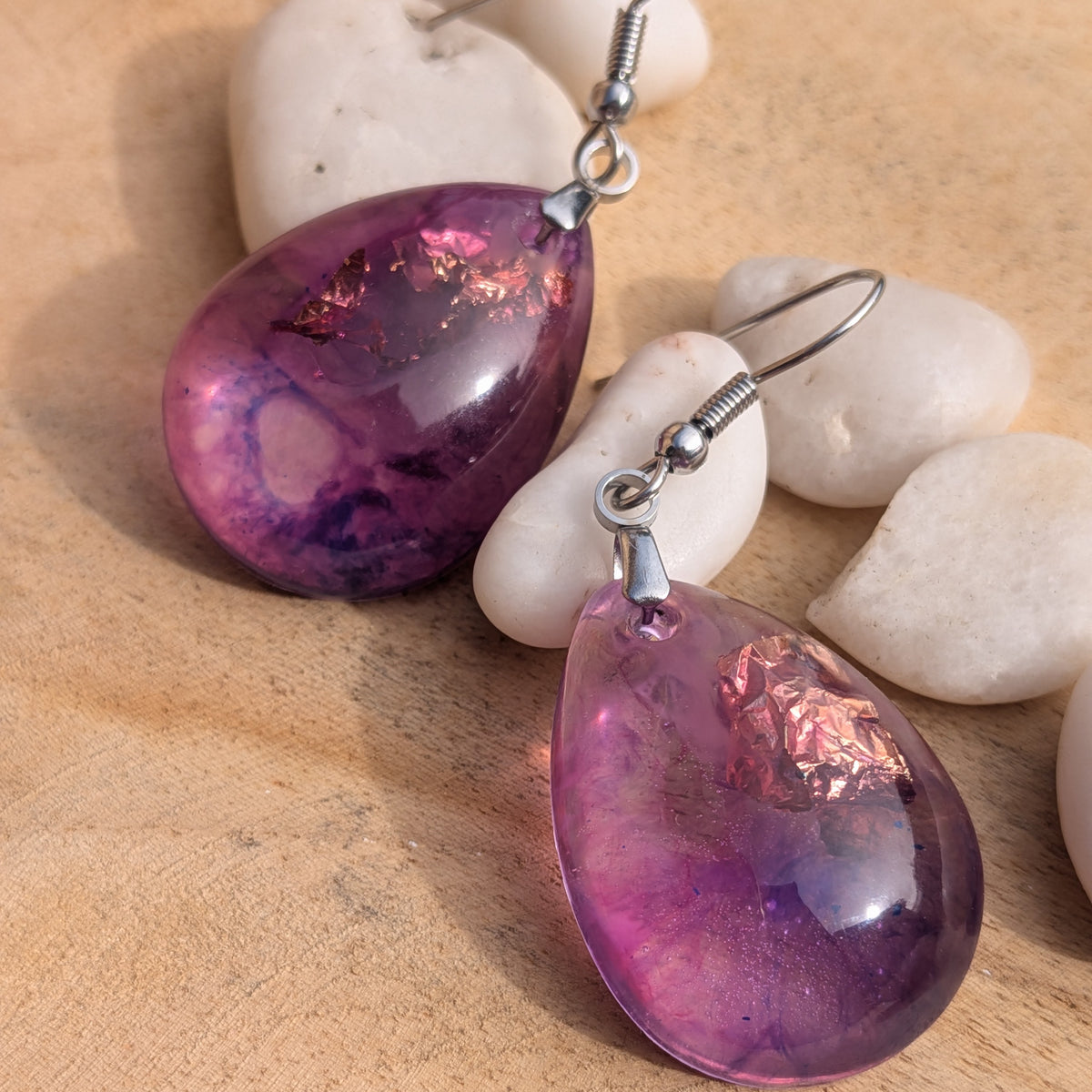 Angled view of violet pink resin earrings laid separately among white stones, EL-Creations