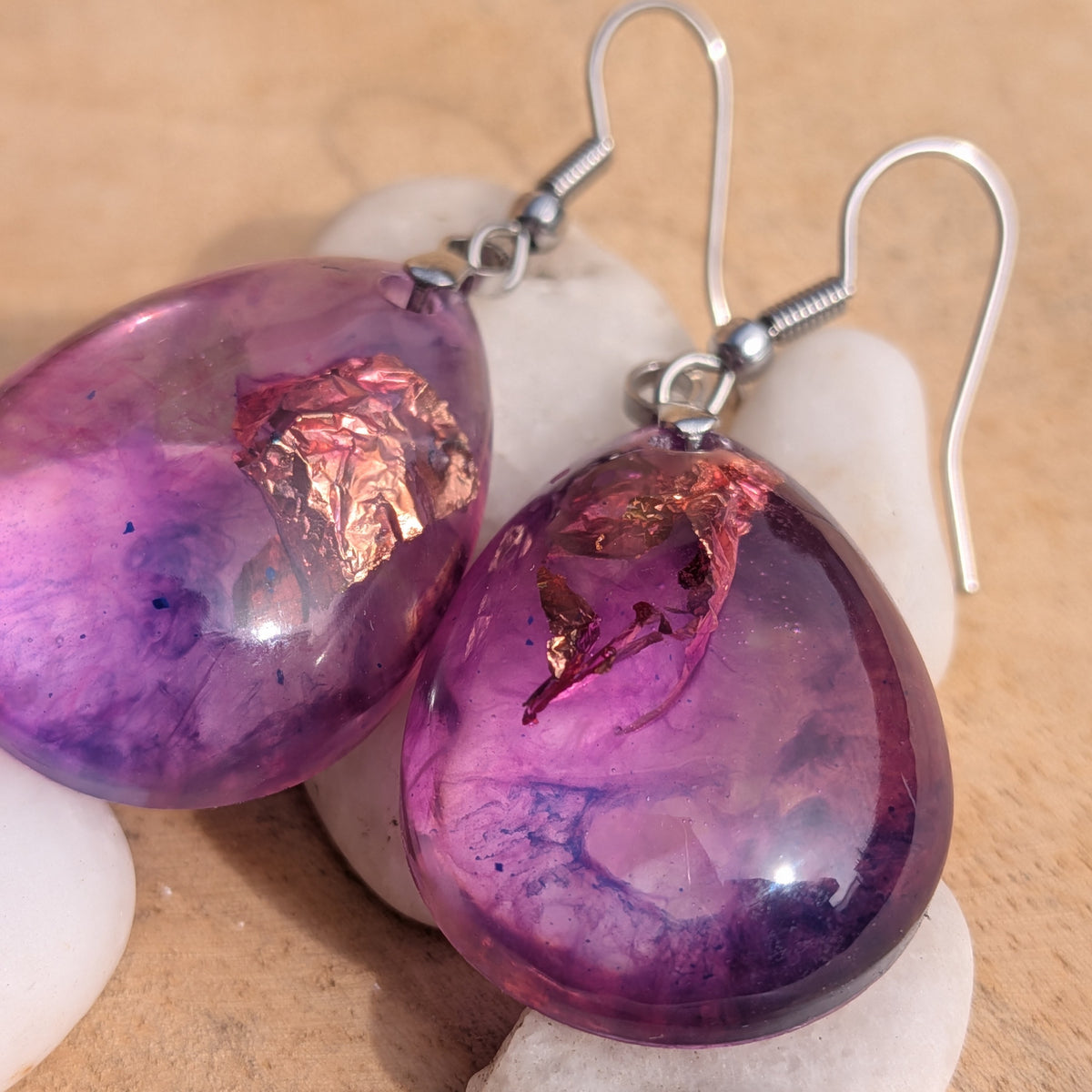 Macro view of violet pink resin drops and stainless steel hooks beside white stones, EL-Creations