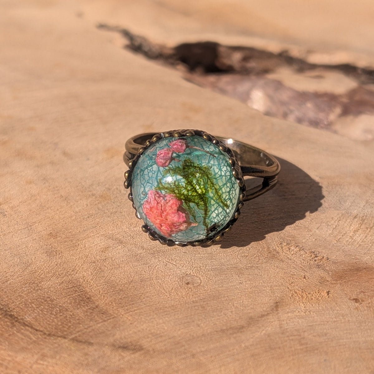 Adjustable botanical resin ring with pink baby’s breath, green moss and blue-green skeleton leaf in a rustic brown setting on wood, EL-Creations