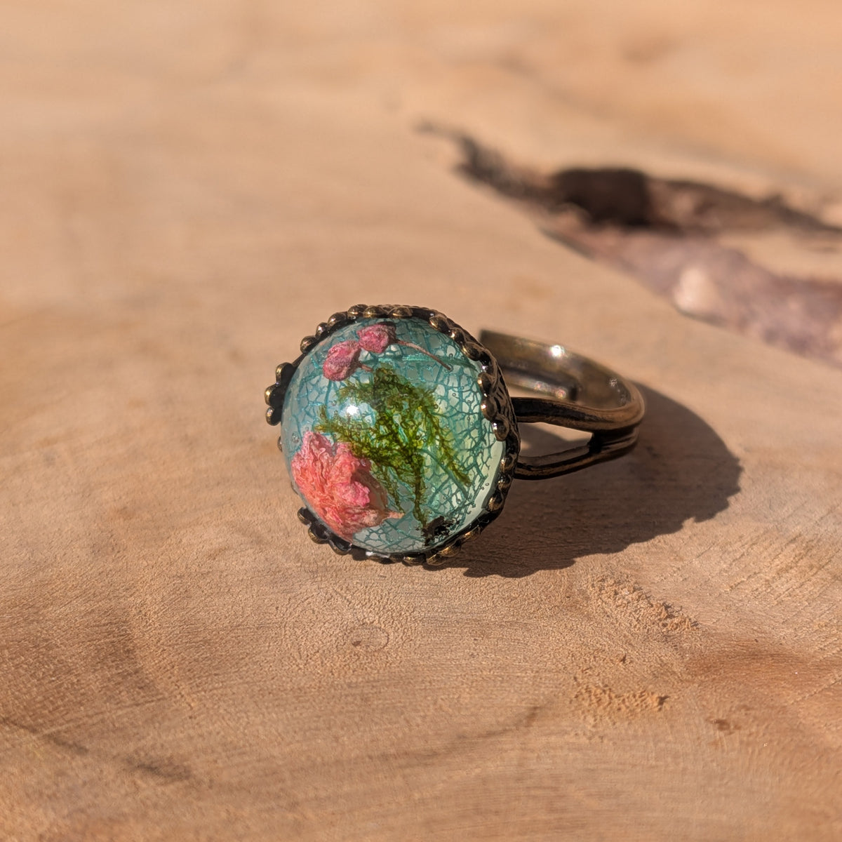 Front view of a botanical resin ring with pink flowers and green moss over a blue-green skeleton leaf in a decorative brown setting, EL-Creations