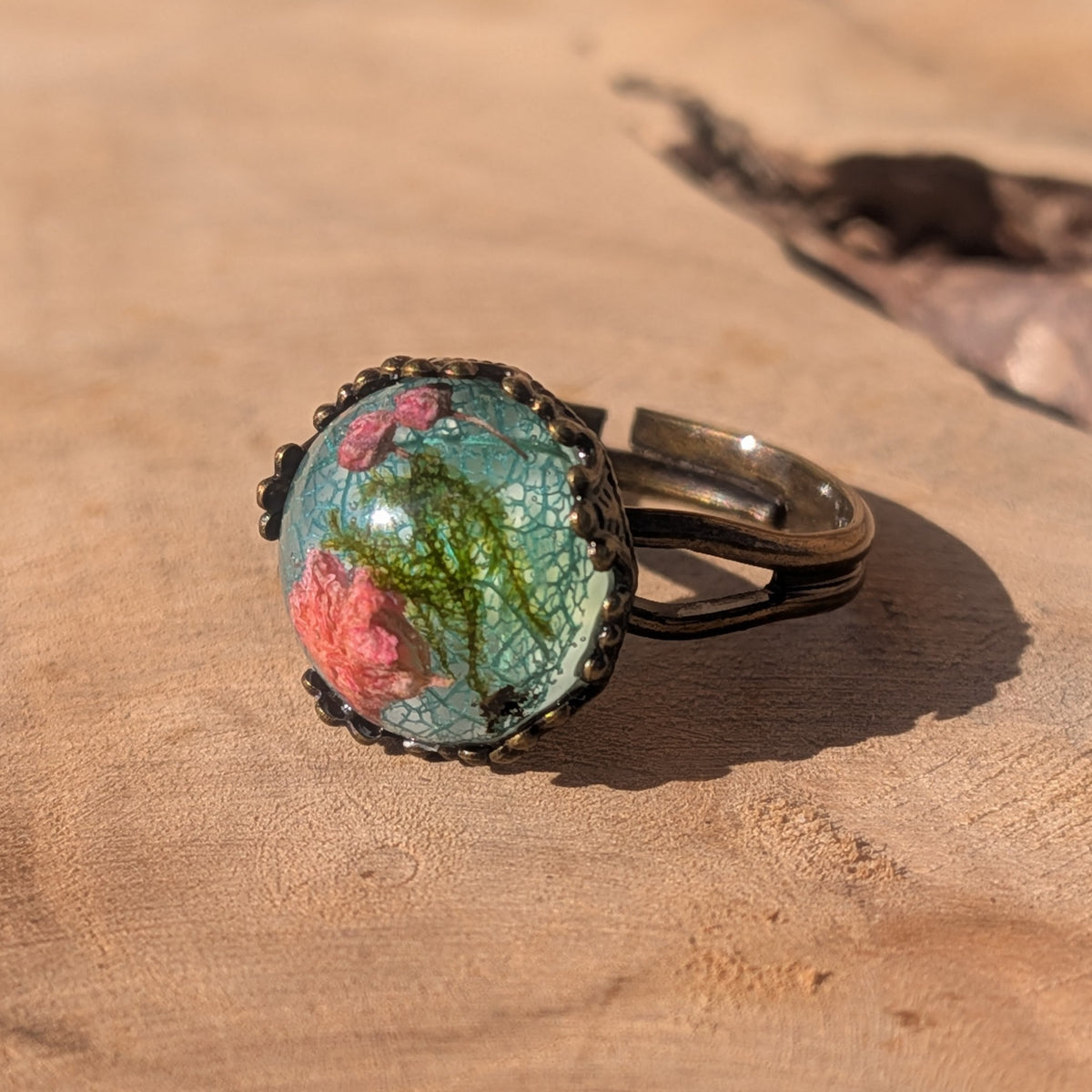 Angled view of a pink baby’s breath and moss resin ring, showing its open adjustable brown alloy band on sunlit wood, EL-Creations