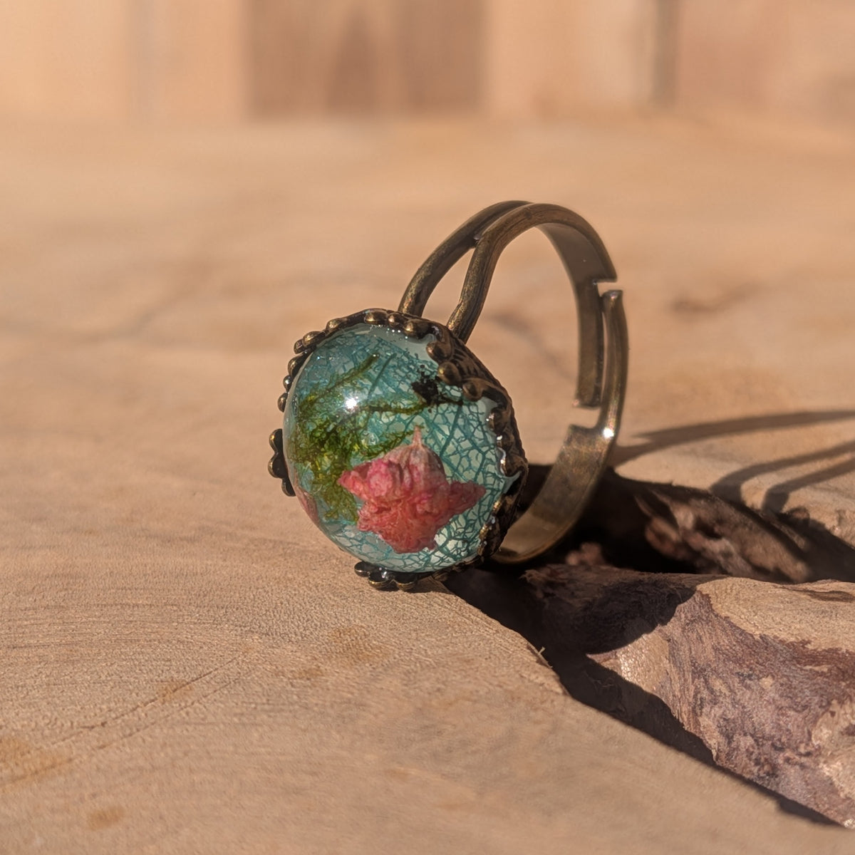 Botanical resin ring standing upright on wood, showing its adjustable band and domed centre with pink baby’s breath and moss, EL-Creations