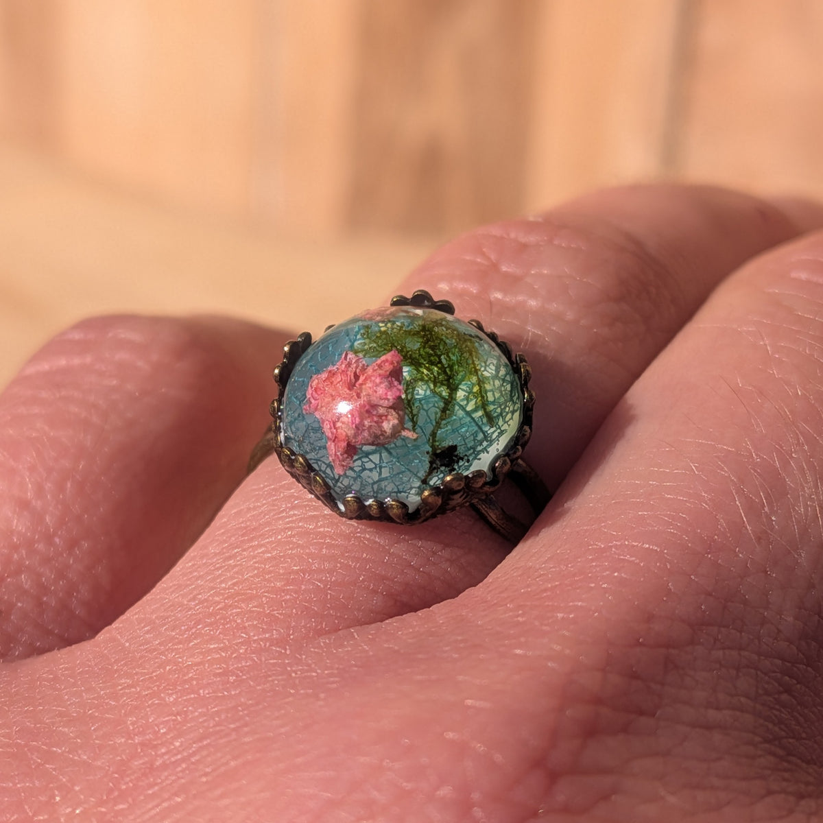 Macro close-up of an adjustable resin ring worn on a finger, showing pink baby’s breath, moss and fine skeleton leaf veins, EL-Creations