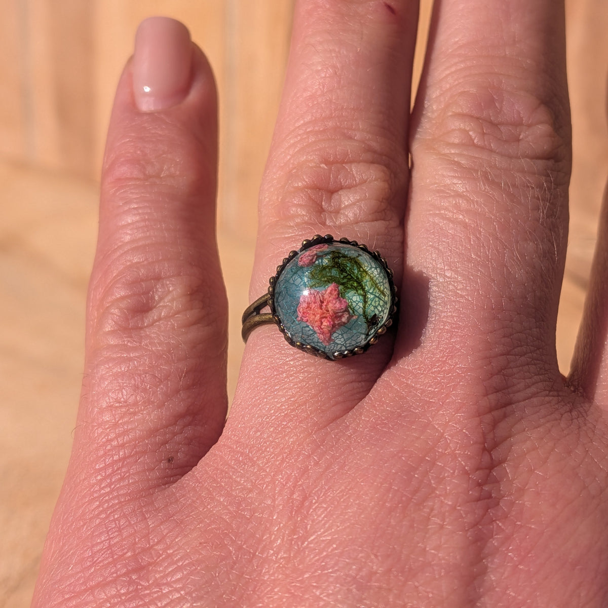 Adjustable botanical resin ring worn on an outstretched hand, showing its pink flowers, green moss and blue-green leaf background, EL-Creations