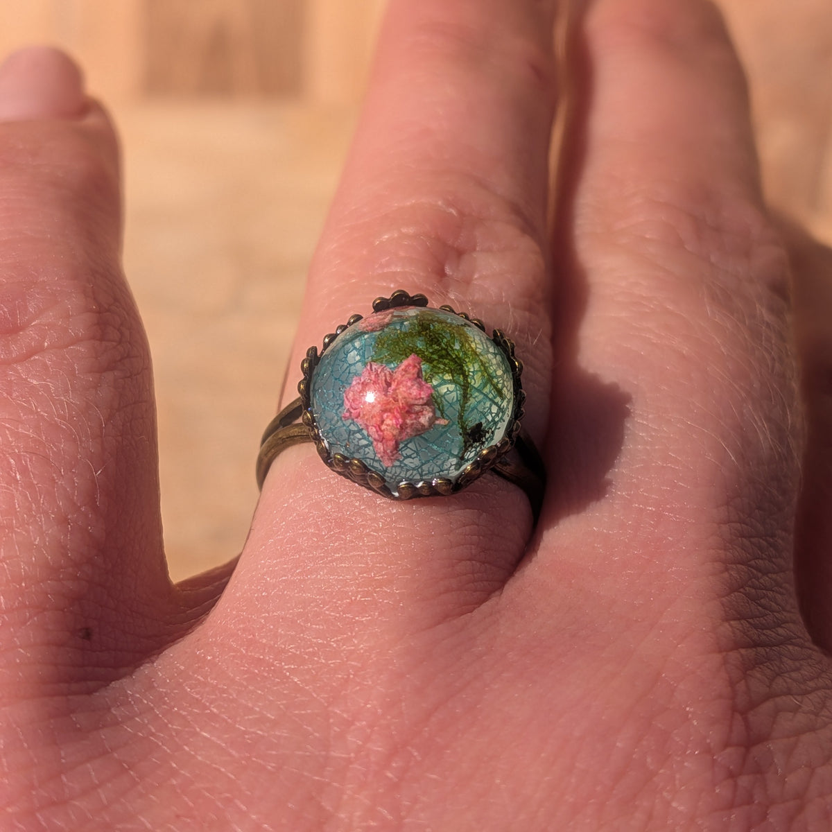 Close-up front view of a pink baby’s breath resin ring worn on a finger in sunlight, framed by a decorative rustic brown border, EL-Creations