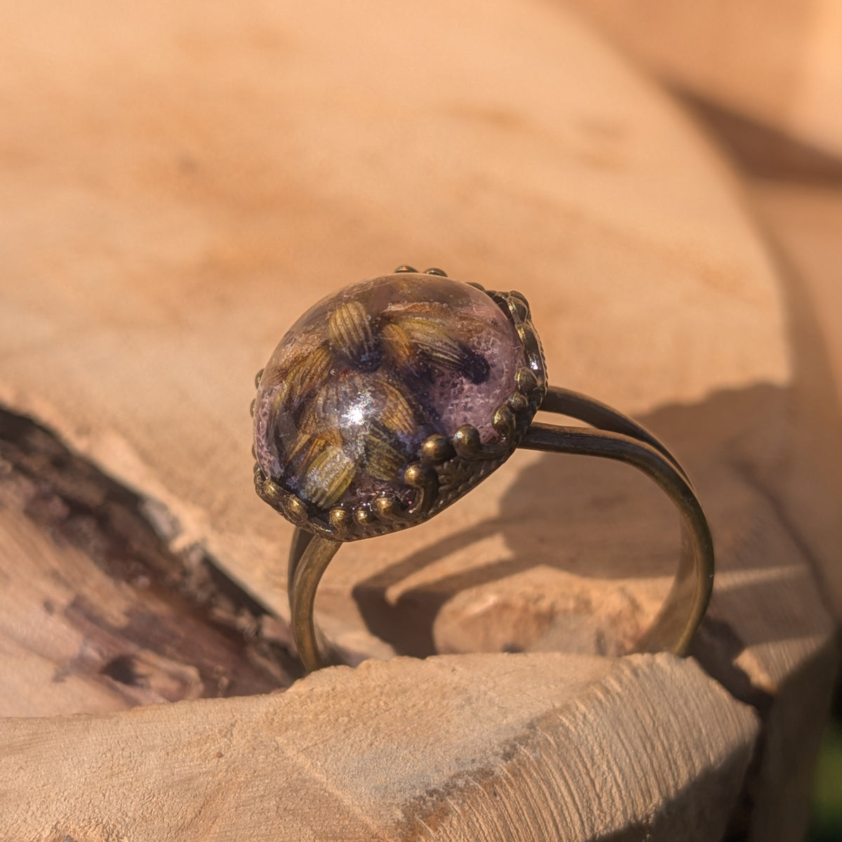 Side view of a lavender and pink leaf resin ring standing upright on wood, showing its domed cabochon and brown alloy band, EL-Creations