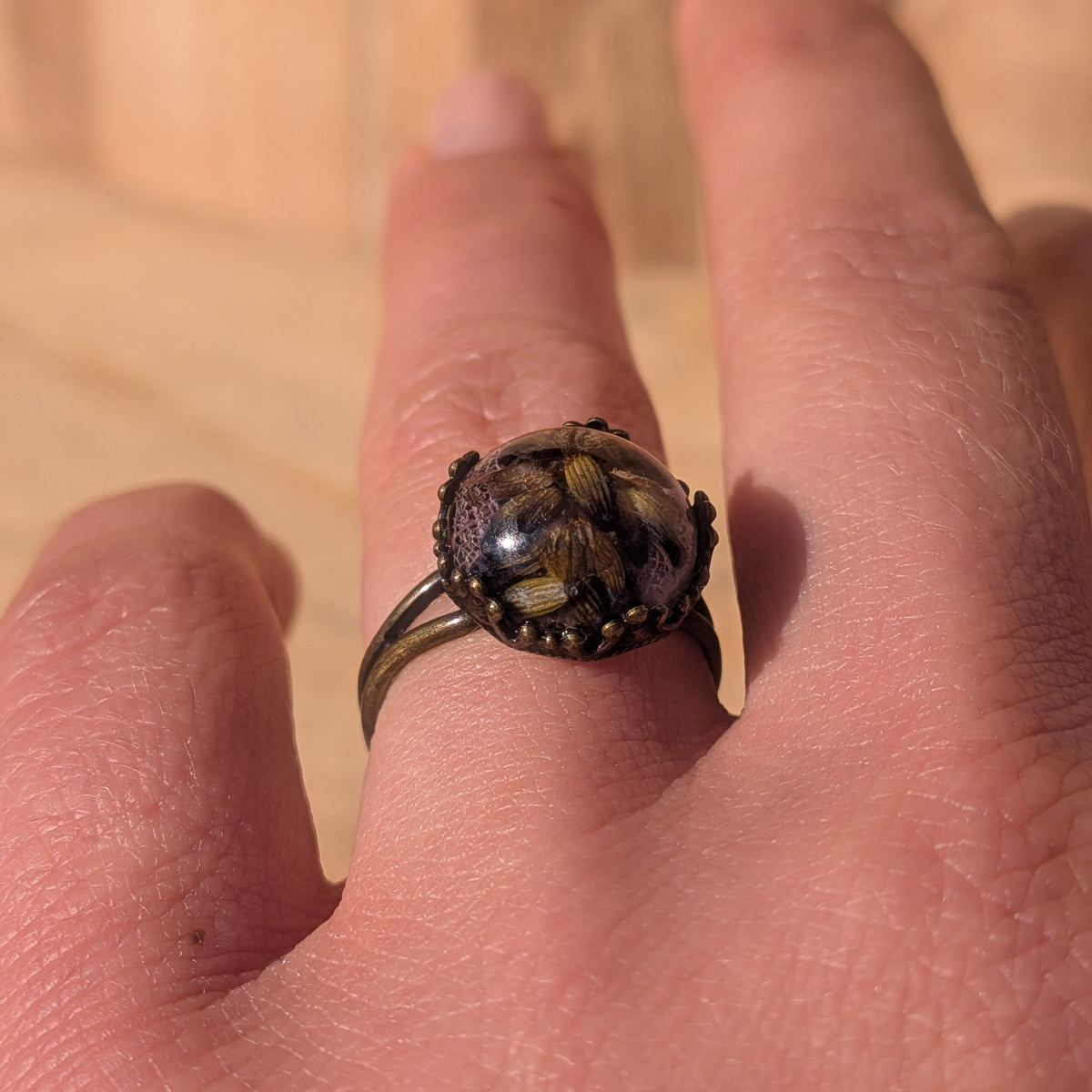 Angled view of an adjustable lavender resin ring worn on a finger, showing its rustic brown split band and botanical cabochon, EL-Creations