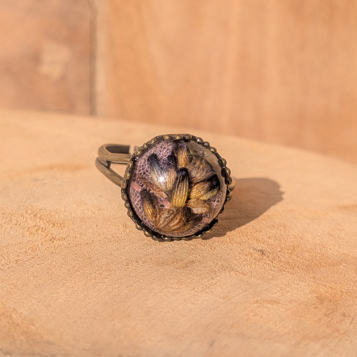 Adjustable resin ring with lavender and pink skeleton leaf in a decorative rustic brown setting on sunlit wood, EL-Creations