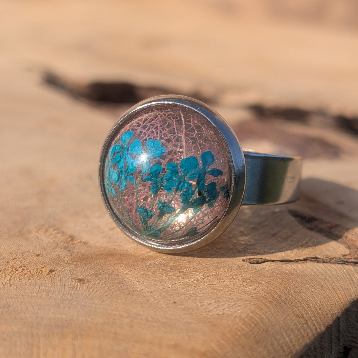 Resin ring with blue Queen Anne’s lace and pink skeleton leaf in a stainless steel setting on sunlit wood, EL-Creations