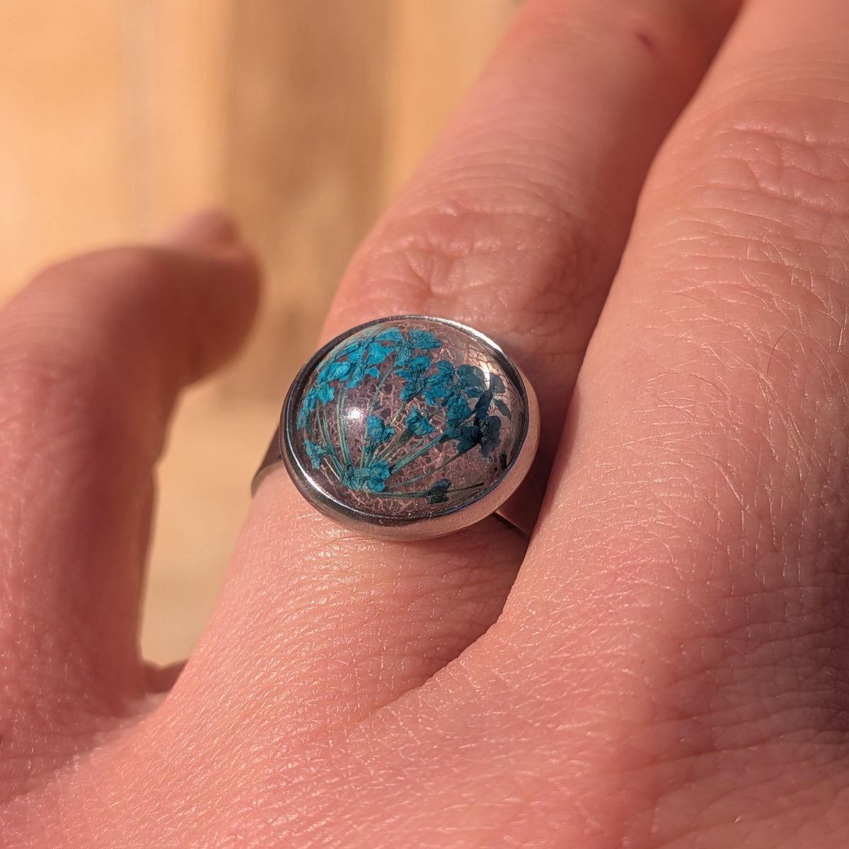 Close-up of a blue Queen Anne’s lace and pink skeleton leaf resin ring worn on a finger in sunlight, EL-Creations