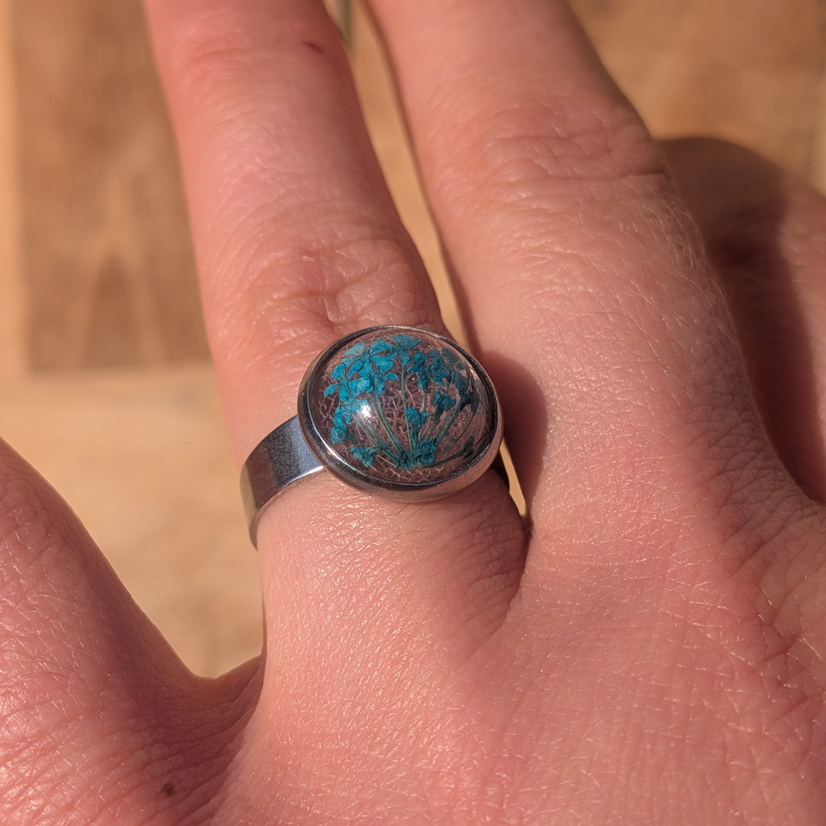 Adjustable botanical resin ring worn on a finger at an angle, showing its wide silver-coloured stainless steel band, EL-Creations