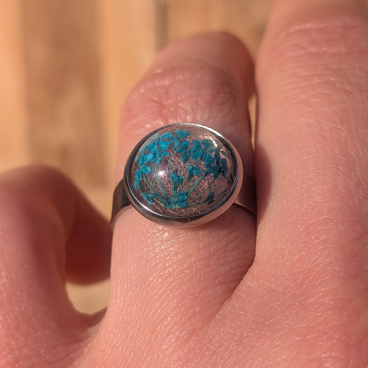 Front macro view of blue Queen Anne’s lace flowers and fine pink leaf veins preserved inside a resin ring worn on a finger, EL-Creations
